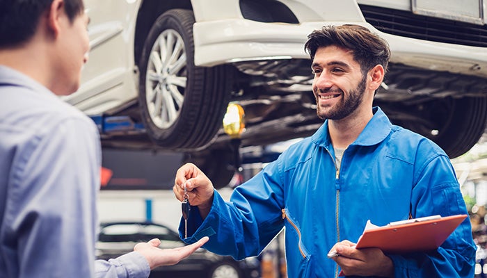 A certified technician has finished servicing a car raised high behind him is giving the keys to its owner.